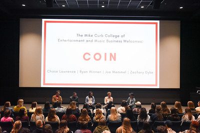 The band COIN sitting in front of an audience, with a background that says "COIN" and lists the members' names