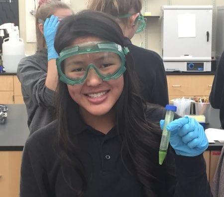 girl, wearing safety goggles, smiling and holding a test tube