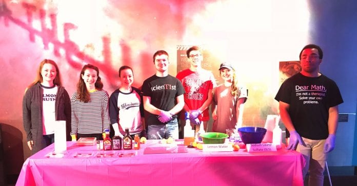 Students hosting the Adventure Science Center experience for children stand behind their experiment table