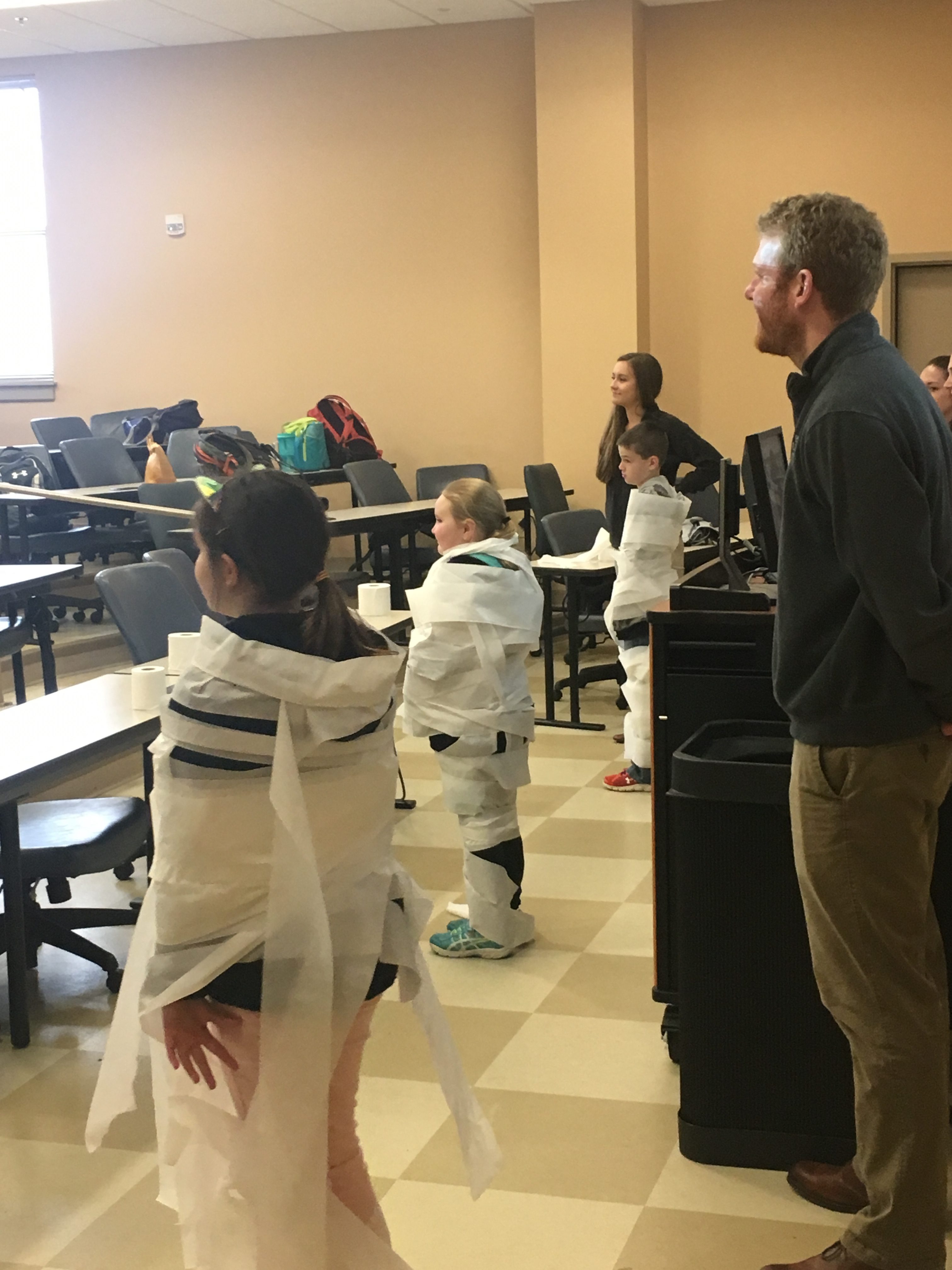 three fourth graders, wrapped in toilet paper, standing with a professor