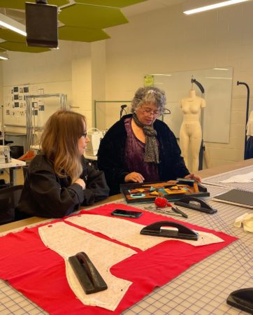 Artist Kateri Pomeroy and fashion student Kat Westmoreland sit at a drafting table and look at Pomeroy mixed media art piece.