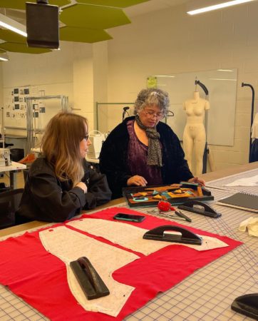 Artist Kateri Pomeroy and fashion student Kat Westmoreland sit at a drafting table and look at Pomeroy mixed media art piece. 