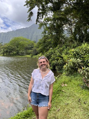 Mia Coutts stands next to the water with a backdrop of a mountain in Hawaii